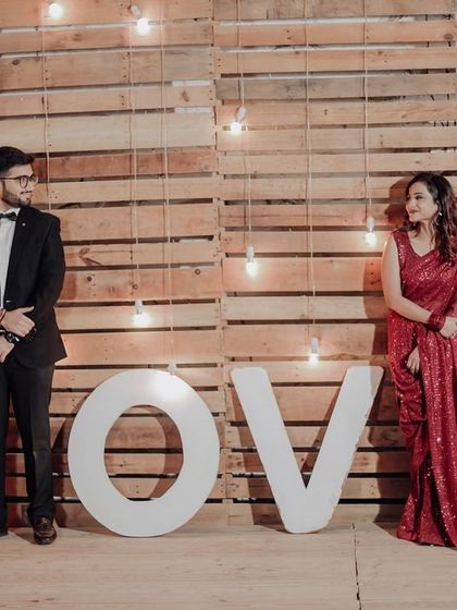 A stylish couple poses with our large 'LOVE' sign against a rustic wooden pallet wall with hanging bulbs. This setup is great for modern and chic pre-wedding or anniversary photos.