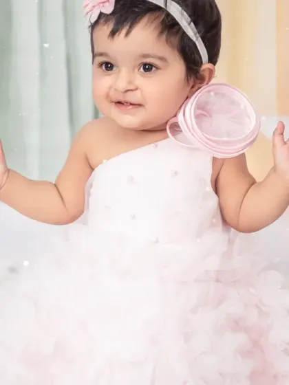 A baby girl in a beautiful white dress, surrounded by a dreamy pastel rainbow and cloud-themed set for her first birthday.