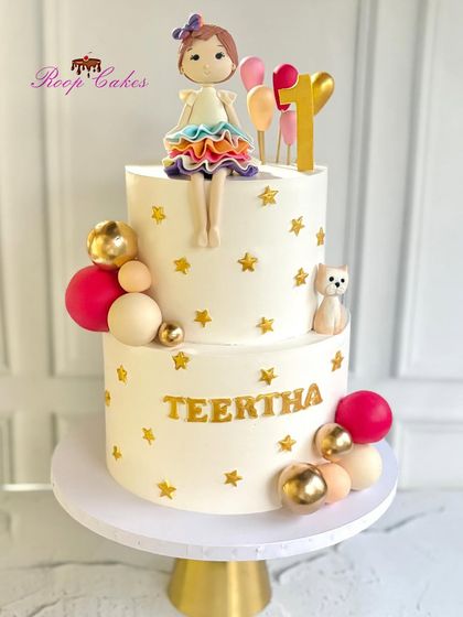 A celebratory two-tier cake for Teertha's first birthday. The design is decorated with gold stars, colorful spheres, and a handcrafted doll topper holding balloons.