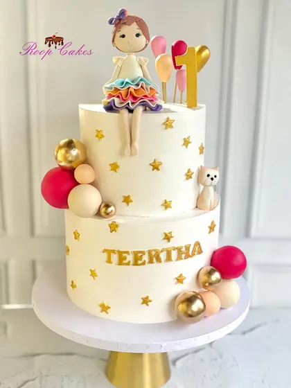 A sweet celebration for a big milestone. This charming two-tier cake for a first birthday is complete with a fondant doll, balloons, and golden stars.