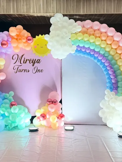 A cheerful rainbow themed first birthday. We created a large balloon arch in pastel rainbow colors, complete with clouds and a smiling sun, against a simple pink backdrop.