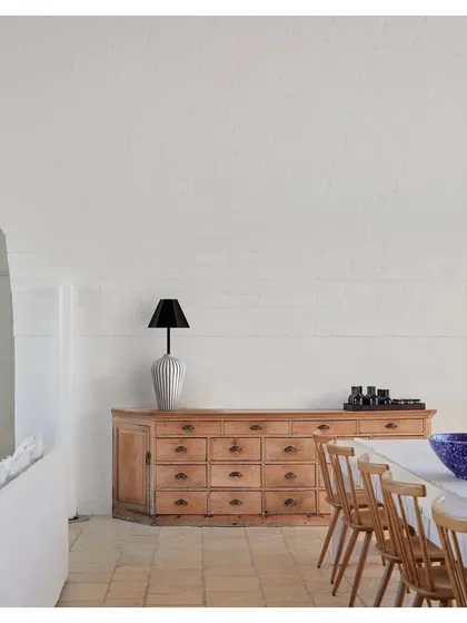 A table lamp from the 'Bernard and friends' collection sits atop a rustic wooden credenza. The lamp's simple, elegant form and natural wood texture bring a sense of calm and craftsmanship to the dining space.