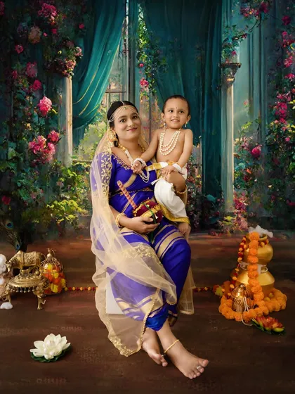 A mother and child portrait set against a magical, floral backdrop. The mother's blue and gold saree complements the baby's traditional Krishna attire perfectly.
