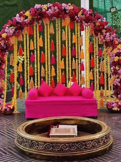 This vibrant Mehndi seating arrangement features a bright pink sofa set against a backdrop of hanging tassels and a beautiful floral arch. The antique-finish urli in the front adds a traditional touch.
