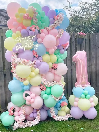 A dreamy pastel balloon wall for an outdoor first birthday celebration. This setup features a 'Happy Birthday' sign, delicate floral accents, and a large pink foil number '1' balloon.