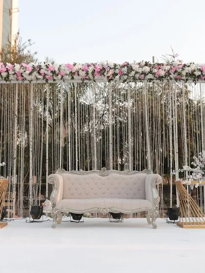 A chic and modern stage for a day wedding, with a backdrop of shimmering silver strings and a lush floral border. The classic white sofa completes the look.