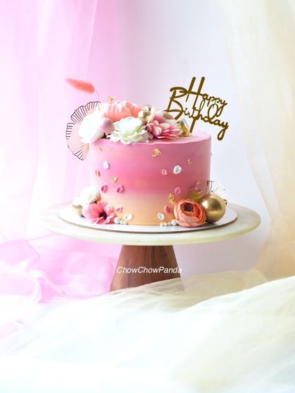 An elegant pink ombre cake decorated with a mix of artificial flowers, gold spheres, and edible gold leaf. This Choco Vanilla cake is perfect for a mom's birthday or any special occasion.
