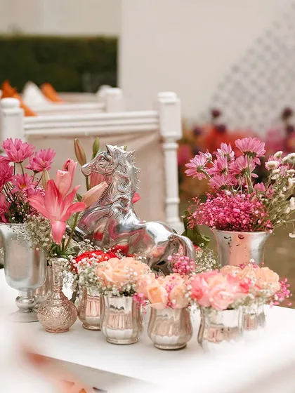 A table centerpiece for a Haldi ceremony, featuring pink flowers and a silver horse figurine. The mix of vibrant colors and metallic accents creates a modern and chic look.