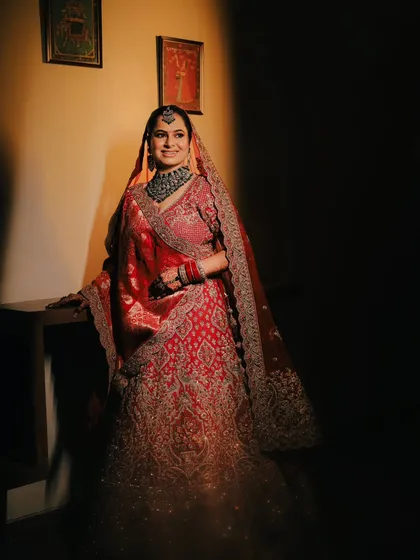 A classic bridal portrait using shadow and light. The bride's stunning red lehenga and intricate jewelry are highlighted as she smiles, creating a timeless image.
