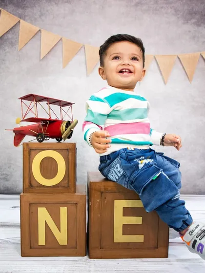 A joyful baby boy sitting on 'ONE' blocks with a toy airplane, set against a rustic studio backdrop.