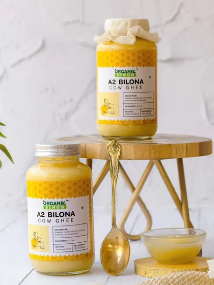 Two jars of A2 Bilona Cow Ghee are styled in a bright, airy setting on a small wooden stool. The clean background and touch of greenery give this shot a fresh, modern, and healthy feel.