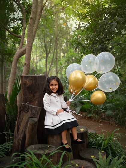 A portrait of a little girl holding balloons, sitting on a tree stump in a beautiful wooded area. The mix of formal wear and a natural setting creates a lovely contrast.