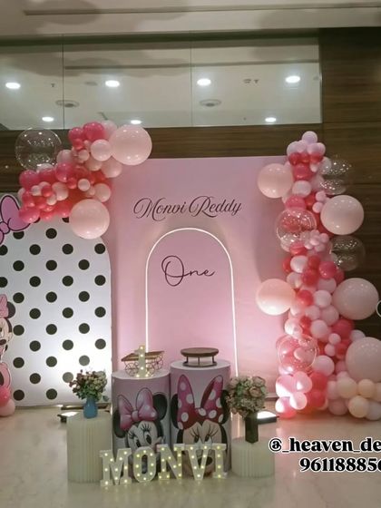 An elegant Minnie Mouse first birthday setup in pink and white. The decor features a modern arched backdrop with neon light accents, Minnie Mouse balloon columns, and custom-printed plinths.