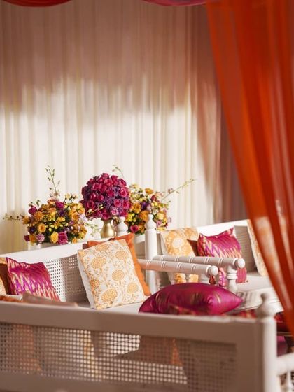 An elegant seating area with white traditional benches, adorned with custom-printed cushions in shades of orange and pink, set against a backdrop of colorful drapes.