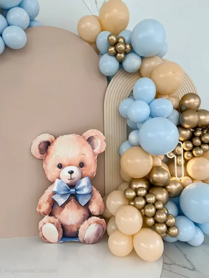 A close-up of a teddy bear-themed backdrop. A cute teddy bear cutout sits beside a balloon garland of soft blue, beige, and chrome gold balloons.