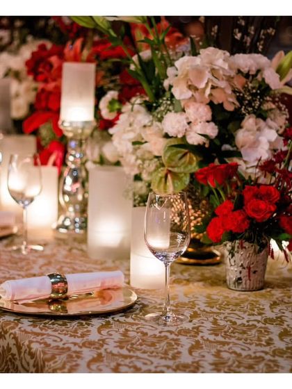 A close up of a formal table setting with red and white floral decor. The rich brocade tablecloth and gleaming glassware create a sense of classic opulence, ideal for a festive wedding dinner.