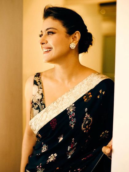 A candid, joyful moment captured during a photoshoot. Kajol's smile radiates in this elegant black saree with a detailed floral border.