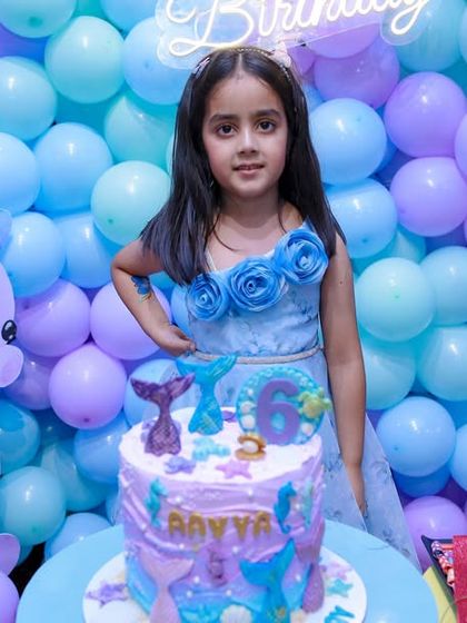 The birthday girl proudly stands with her mermaid-themed cake against a backdrop of purple and blue balloons, creating a perfect color-coordinated photo.