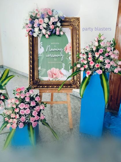 A personalized welcome sign for a naming ceremony, framed in ornate gold and topped with a lush arrangement of pink and purple flowers. The surrounding floral stands continue the color theme, creating a cohesive and inviting entrance.