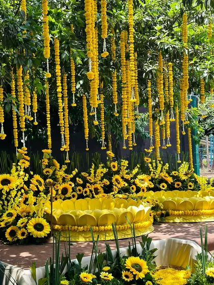A wider view of the outdoor Haldi seating, with two large yellow lotus props and marigold garlands hanging from the trees above.