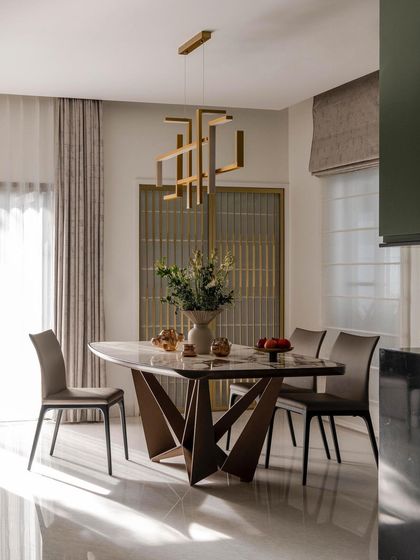 The dining area in the 'ARR' residence features a sculptural table base and a geometric brass chandelier. Sunlight streams in, highlighting the polished marble floors and creating a bright, elegant space for formal dining.