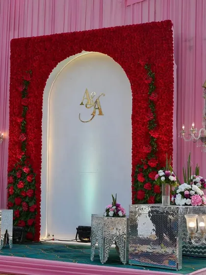 A modern and glamorous stage setup. The red floral arch against a white backdrop, paired with mirrored tables, creates a chic and sophisticated look with a pop of color.