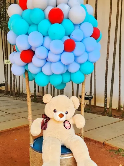 A creative and fun decor piece for a baby shower or first birthday. This prop features a large teddy bear sitting in a basket attached to a "hot air balloon" made entirely of blue, white, and red balloons.