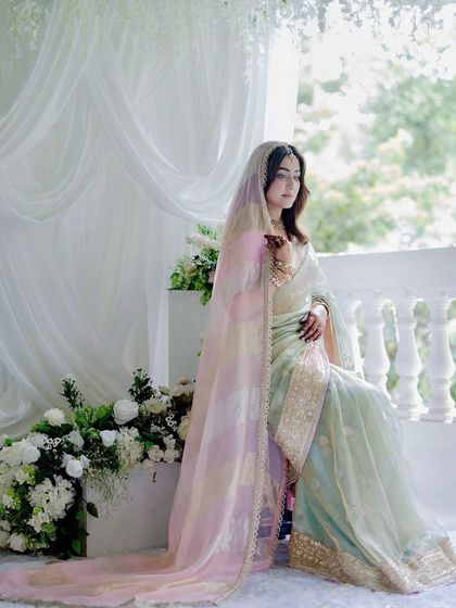 The bride seated by a classic white balustrade, with lush floral arrangements and soft drapes creating a timeless and romantic scene.