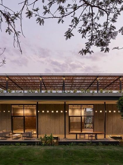 The building's facade at dusk, framed by native trees. The combination of the thatched upper roof, large glass openings, and warm interior lighting creates a sense of transparency, blurring the boundary between the indoor spaces and the surrounding garden.