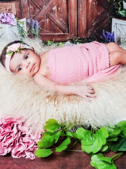 An awake baby girl rests on a miniature bed surrounded by pink flowers. Her calm expression and direct gaze make for a beautiful portrait.