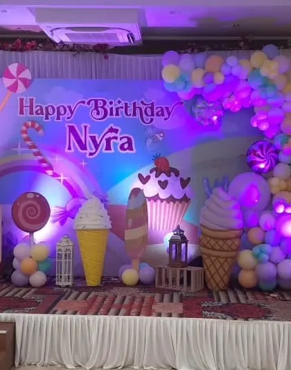 A stage setup for Nyra's Candyland birthday, featuring a custom printed backdrop with a candy road, large ice cream cone props, and a pastel balloon arch.