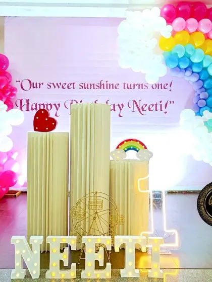 A bright and cheerful rainbow-themed first birthday. The centerpiece is a large balloon rainbow arch over a personalized backdrop, complemented by pink balloon columns and light-up marquee letters for the name.
