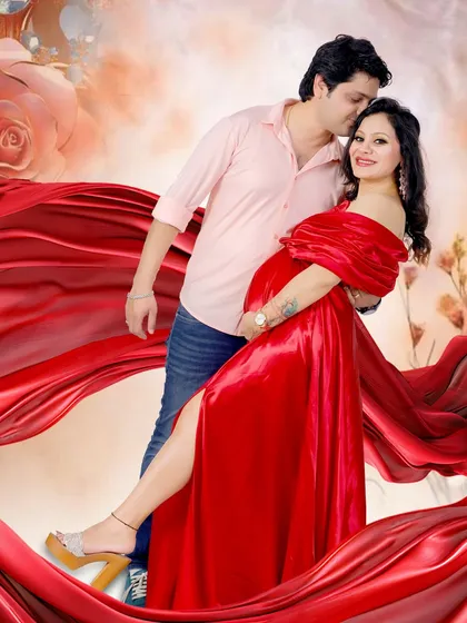 A glamorous studio portrait of an expecting couple. The vibrant red satin gown with its flowing fabric creates a dramatic and romantic feel.