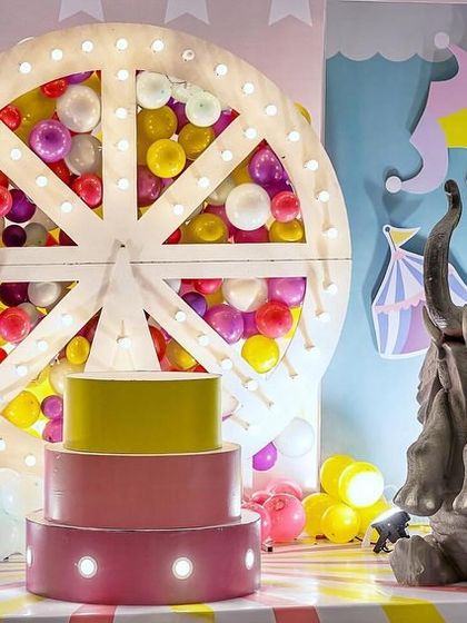 A detailed shot of the carnival cake stage, featuring a three-tier faux cake stand with lights and a playful elephant prop. The lit Ferris wheel in the background is filled with colorful balloons.