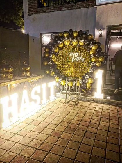 A first birthday party setup at an outdoor venue, featuring a circular balloon arch and large illuminated letters spelling the name 'HASIT'.