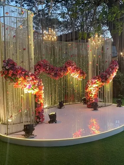 A unique and modern stage backdrop featuring a wavy structure adorned with crystals and a flowing arrangement of pink flowers. The spotlights from below highlight the texture and shape of the design.