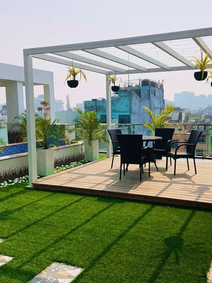 A rooftop escape in Dwarka. This design uses a clean, white metal pergola to create a shaded dining area. The space is defined by a wooden deck, artificial turf, and planters with palms, making it a perfect spot for enjoying the city view.