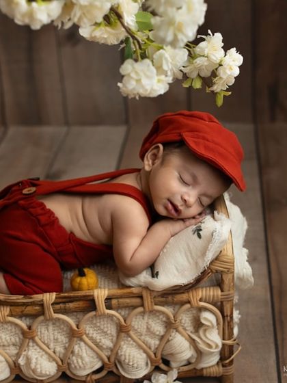 A sweet newborn moment in a red outfit, sleeping on a tiny pillow in a wicker bed. The warm tones and cozy setup make this a truly heartwarming image.