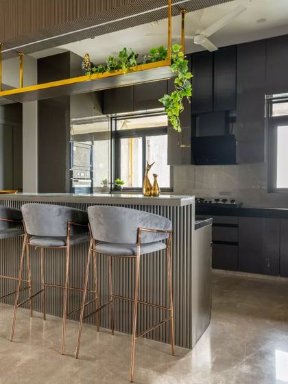 The high-rise island counter in this open kitchen is a commanding structure that invites both culinary creativity and social interaction. A suspended brass shelf adorned with cascading greenery offers a subtle touch of organic warmth, softening the kitchen’s sharp lines.