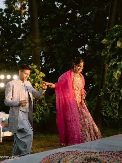 He leads her towards the celebration, a happy, candid moment just before their engagement. The warm light and her bright pink lehenga create a vibrant, joyful scene.