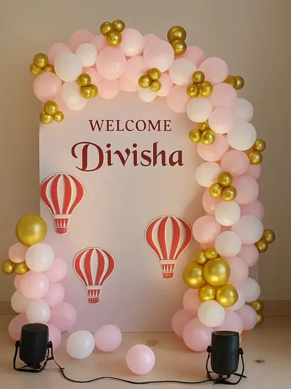 A beautiful and classic welcome party for a newborn baby girl. This setup features a pink, white, and gold balloon arch over a custom backdrop with a hot-air balloon theme, creating a sweet photo spot.