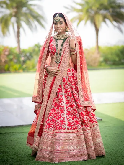 A full-length portrait of a bride looking absolutely majestic. The soft glam makeup with defined eyes stands out beautifully against her ornate red and gold lehenga, creating a truly royal vision.