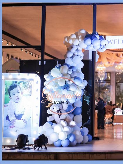 A balloon sculpture at the entrance, featuring a teddy bear surrounded by a cascade of blue and white balloons next to a personalized, illuminated photo frame of the baby.