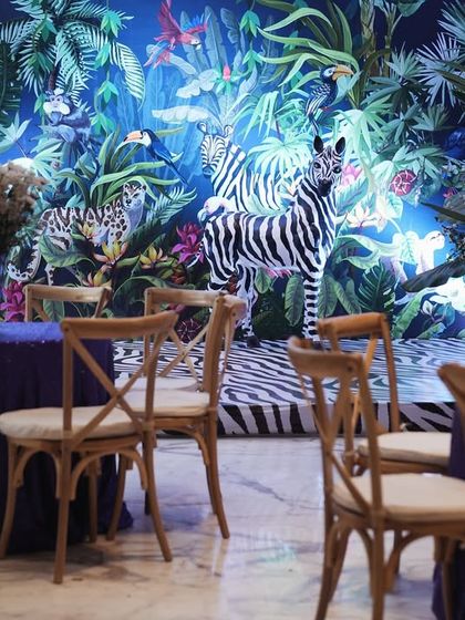 A seating area within the jungle-themed event. The backdrop is a beautiful wallpaper featuring zebras and tropical birds, with rustic wooden chairs and deep blue tablecloths completing the immersive experience.
