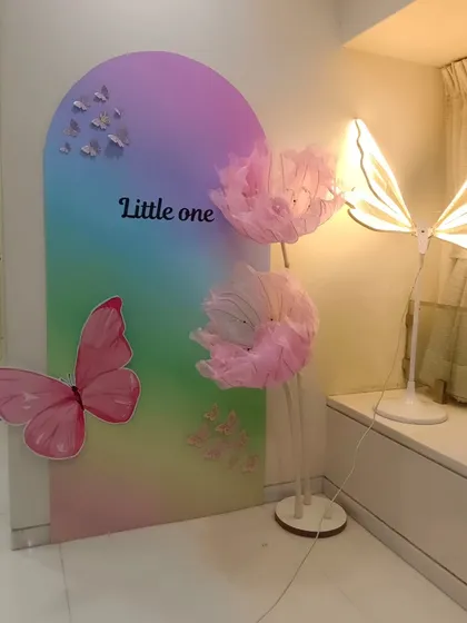 A modern art-style backdrop for a 'Little One' celebration, featuring a rainbow gradient panel, a large pink butterfly, and delicate light-up props.