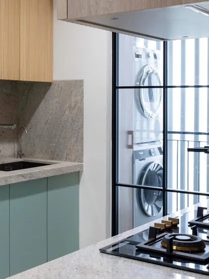 A glass door separates the main kitchen from the utility balcony where the laundry machines are stacked. This visually connects the spaces while containing any noise, creating a smart and practical layout.