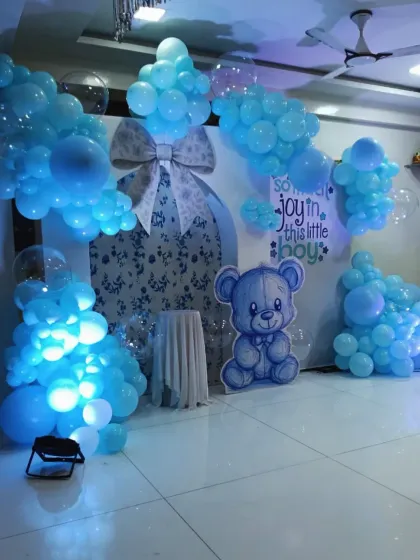 The full view of the gift box and teddy bear baby shower decoration. The scene is filled out with clusters of blue and transparent balloons, with spotlights to highlight the setup.