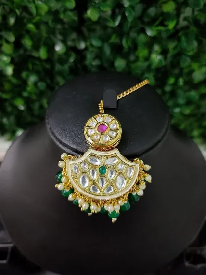 A delicate maang tikka with a small crescent design, featuring kundan work and green and pink stones.