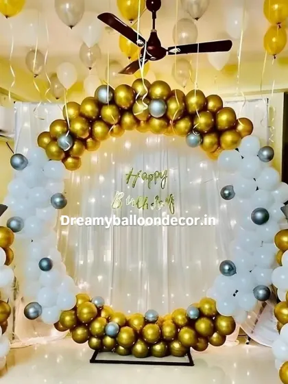 An elegant gold and white themed birthday backdrop. This home setup features a full balloon ring with chrome gold and white balloons, star-shaped foil balloons, and a sheer curtain with fairy lights.