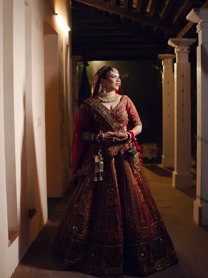 A full-length portrait of the bride in a richly detailed red lehenga. The soft lighting in the corridor highlights the exquisite craftsmanship of her wedding attire.